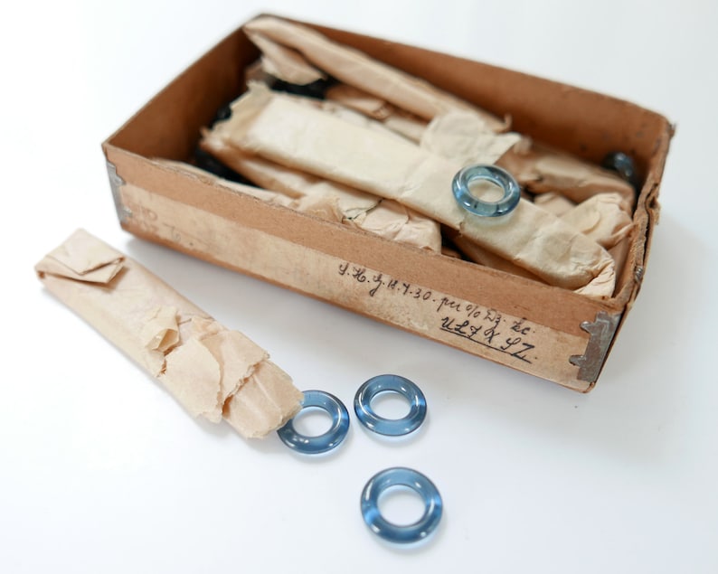 May include: A box of blue glass rings, some are wrapped in brown paper, and some are loose on a white surface. The box is labelled with handwritten text: "y.z.g.y.h. 30. per % 5g. zc. u2.4 x 97."