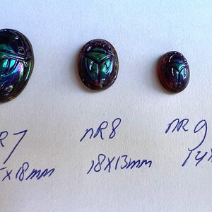 May include: Three iridescent glass scarab beetle cabochons in different sizes. The largest measures 2.5 inches by 1.8 inches, the middle measures 1.8 inches by 1.3 inches, and the smallest measures 1.4 inches by 1.0 inches.