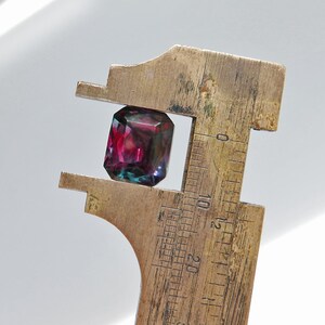 May include: A close-up of a dark purple and green gemstone being measured with a brass caliper. The gemstone is approximately 10 millimeters in diameter.