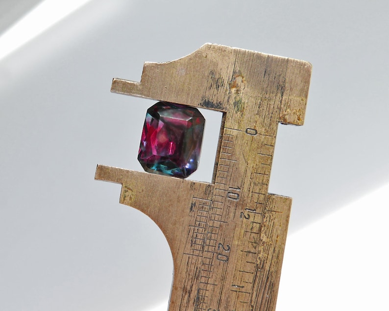 May include: A close-up of a dark purple and green gemstone being measured with a brass caliper. The gemstone is approximately 10 millimeters in diameter.