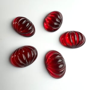 Pode incluir: Cinco cabochões de vidro vermelho com textura nervurada. Os cabochões são de forma oval e têm um acabamento brilhante.