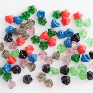 Puede incluir: Surtido de cuentas de vidrio de colores en varias formas, incluyendo cuentas en forma de estrella en vidrio rojo, azul, verde y transparente. Las cuentas están esparcidas sobre una superficie blanca.