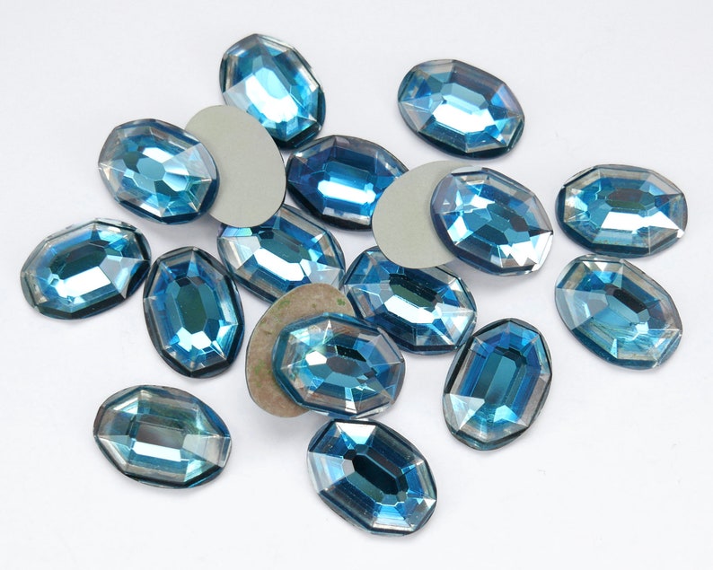 May include: A collection of oval-shaped, faceted, blue rhinestones. The rhinestones have a clear, reflective surface and are scattered on a white background.