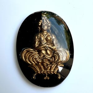 May include: An oval black resin pendant with a gold-coloured seated Buddha figure. The Buddha is in a meditative pose on a lotus flower. The pendant has a glossy finish, and the Buddha figure has intricate details.