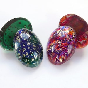 May include: Two pairs of oval-shaped glass cabochons. One pair is green with gold flecks, the other is purple with gold flecks.