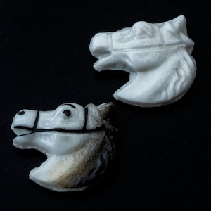 May include: Two horse head figurines. One is solid white, the other is white with gray and black accents. The horse heads are in profile, with detailed features including bridles and facial expressions. The figurines are set against a black background.
