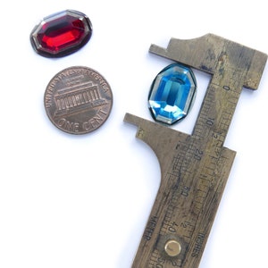 May include: A close-up of a blue and red glass gem stones being measured with a vintage brass caliper. The caliper has a ruler marked in inches and centimeters.  The gem stones are being measured against a penny.