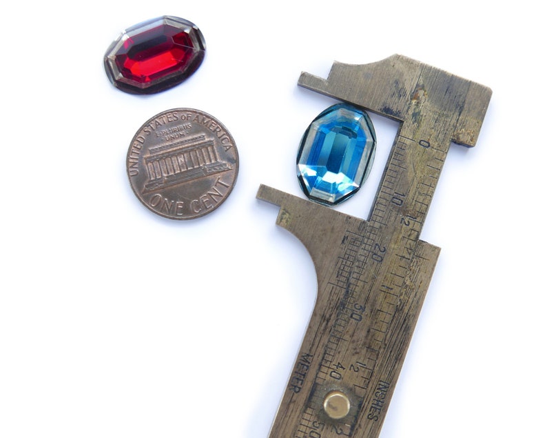 May include: A close-up of a blue and red glass gem stones being measured with a vintage brass caliper. The caliper has a ruler marked in inches and centimeters.  The gem stones are being measured against a penny.