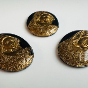 May include: Three vintage-style, round buttons featuring a gold relief of a woman's face and draped fabric, set against a black background. The buttons are arranged on a white surface.