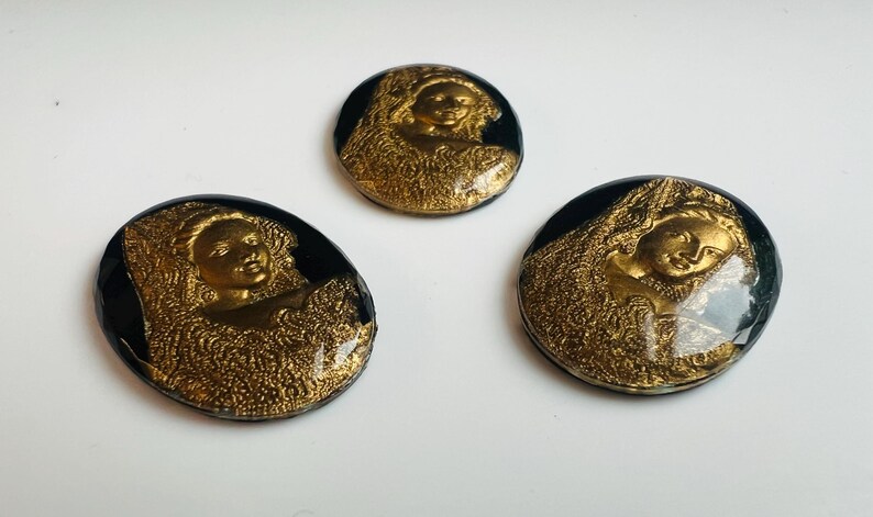 May include: Three vintage-style, round buttons featuring a gold relief of a woman's face and draped fabric, set against a black background. The buttons are arranged on a white surface.