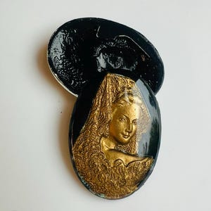 May include: An oval brooch with a gold relief of a woman's face and hair, set against a black background. The brooch has a glossy finish and a vintage aesthetic.
