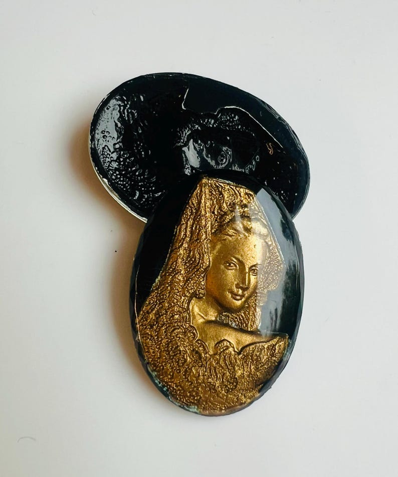 May include: An oval brooch with a gold relief of a woman's face and hair, set against a black background. The brooch has a glossy finish and a vintage aesthetic.