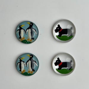 Pode incluir: Quatro botões redondos de vidro com imagens pintadas. Dois botões apresentam um par de pinguins sobre um fundo azul. Dois botões apresentam um cão preto com uma coleira vermelha sobre um fundo verde.