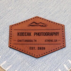 May include: A brown leather patch with the text "Kodiak Photography" in black lettering. The patch also includes the text "Chattanooga, TN" and "Athens, GA" in smaller black lettering. The text "EST. 2020" is also included in black lettering.