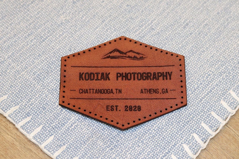 May include: A brown leather patch with the text "Kodiak Photography" in black lettering. The patch also includes the text "Chattanooga, TN" and "Athens, GA" in smaller black lettering. The text "EST. 2020" is also included in black lettering.