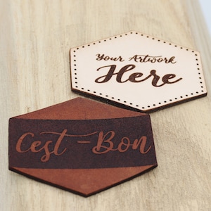 May include: Two hexagonal leather patches with text. The top patch is light brown with the text "Your Artwork Here" in brown. The bottom patch is brown with a dark brown stripe and the text "C'est Bon" in brown.