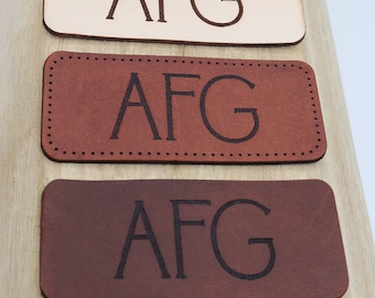 May include: Three leather patches with the letters "AFG" embossed in black. The patches are in different shades of brown, from light to dark.