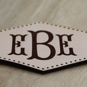 May include: A hexagonal leather patch with a brown monogram of the letters "EBE" in a decorative font.