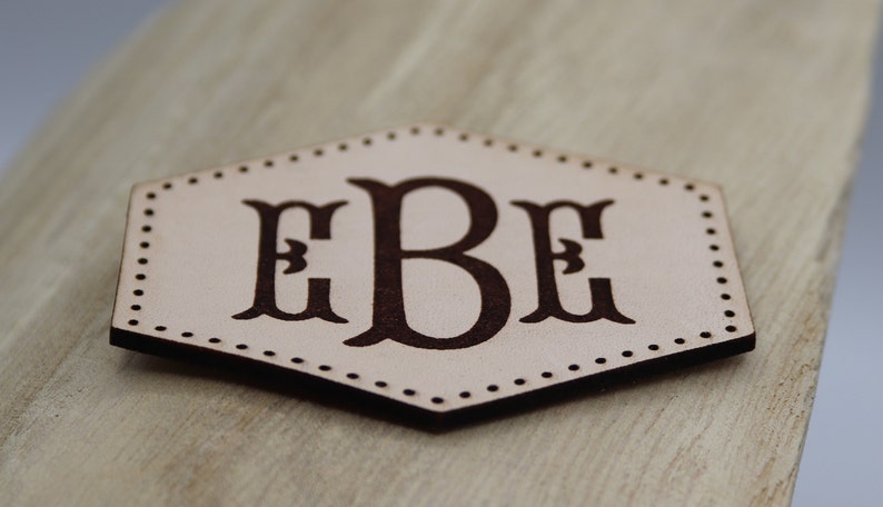 May include: A hexagonal leather patch with a brown monogram of the letters "EBE" in a decorative font.