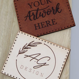 May include: Two leather labels with stitched edges. The top label is brown and says "YOUR Artwork HERE". The bottom label is cream colored and has a circle with a floral design and the text "A.G. DESIGN".