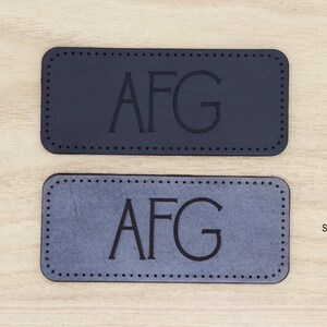 May include: Two leather labels with the initials "AFG" embossed in black. The top label is black and the bottom label is slate gray.