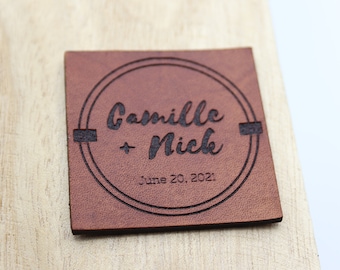 May include: A brown leather square tag with a circle design engraved with the text "Camille + Nick" and the date "June 20, 2021".