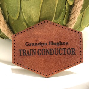 May include: A brown leather tag with a stitched border and the text "Grandpa Hughes TRAIN CONDUCTOR" in black.