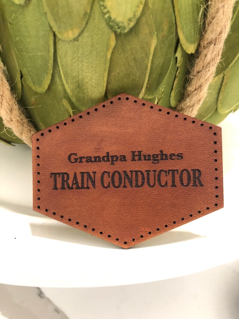 May include: A brown leather tag with a stitched border and the text "Grandpa Hughes TRAIN CONDUCTOR" in black.