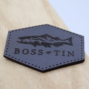 May include: A dark gray leather patch with a fish and mountain design. The text "BOSS TIN" is printed below the fish.