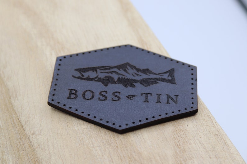 May include: A dark gray leather patch with a fish and mountain design. The text "BOSS TIN" is printed below the fish.