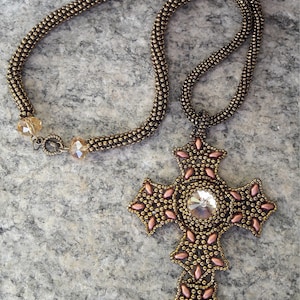 May include: A gold beaded necklace with a large cross pendant. The cross is made of gold beads and has a clear crystal in the center. The necklace is on a gray background.