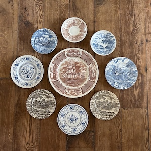 May include: A collection of antique ceramic plates and saucers with various blue and brown transferware designs. The plates feature scenes of landscapes, floral patterns, and a ship. The arrangement is on a wooden surface.