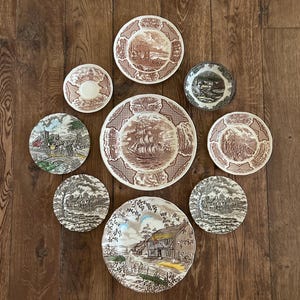 May include: A collection of eight ceramic plates and saucers featuring various transferware designs in brown, green, and sepia tones. The pieces display detailed illustrations of sailing ships, rural landscapes, and architectural scenes with decorative borders.