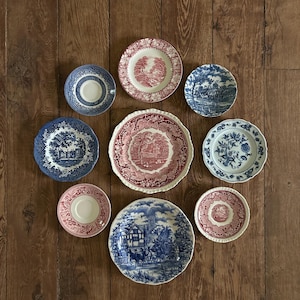 May include: A collection of eight ceramic plates and saucers featuring transferware patterns in shades of blue, white, and red. The pieces display various landscape, architectural, and floral motifs with scalloped or smooth edges in different sizes.