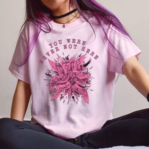 May include: Pale pink t-shirt featuring a pink winged creature graphic and the text "YOU WERE NEVER NOT HERE". The shirt is made of a soft material.