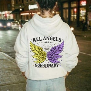 Może przedstawiać: Biała bluza z kapturem z napisem "ALL ANGELS ARE NON-BINARY" i grafiką żółtych i fioletowych skrzydeł. Bluza z kapturem jest noszona przez osobę w jasnoniebieskich dżinsach, stojącą na ulicy miasta w nocy.