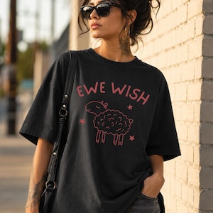 Peut inclure: Un t-shirt noir à manches courtes avec un motif rose représentant un mouton aux yeux fermés et le texte EWE WISH imprimé au-dessus, accompagné de petites étoiles roses entourant l'illustration du mouton sur le devant du vêtement.
