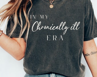 Może przedstawiać: Ciemnoszary t-shirt z krótkim rękawem z białym nadrukiem IN MY Chronically ill ERA. Koszulka ma wykończenie w stylu vintage z efektem sprania oraz okrągły dekolt. Wzór jest wyśrodkowany na klatce piersiowej przy użyciu czcionek szeryfowych i pisanych.