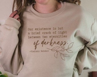 Op de afbeelding: Een beige sweatshirt met een citaat gedrukt op de voorkant: "Ons bestaan is slechts een korte scheur van licht tussen twee eeuwigheden van duisternis." - Vladimir Nabokov. Het citaat is gedrukt in bruin met een klein blaadjesontwerp.
