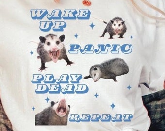 May include: White t-shirt with a graphic of a possum and the text "Wake Up Panic Play Dead Repeat" in blue letters.