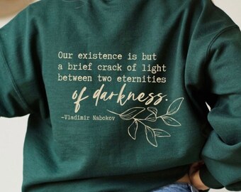 Op de afbeelding: Een donkergroen sweatshirt met een citaat van Vladimir Nabokov gedrukt op de achterkant. Het citaat luidt: "Ons bestaan is slechts een korte scheur van licht tussen twee eeuwigheden van duisternis."