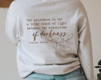 Op de afbeelding: Lichtgrijs sweatshirt met de quote "Our existence is but a brief crack of light between two eternities of darkness" van Vladimir Nabokov. De quote is in bruine tekst met een bladontwerp.