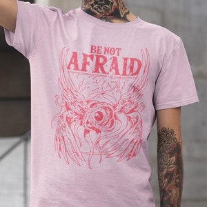 May include: A pink t-shirt with a red graphic print of an eye with wings and the text "BE NOT AFRAID".