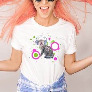 Pu&ograve; includere: T-shirt bianco con una grafica di un gattino tigrato grigio con le cuffie e la bocca spalancata. Il design include cerchi verdi e rosa. La maglietta &egrave; indossata da una persona con i capelli rosa e occhiali da sole.