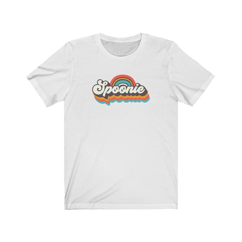 Op de afbeelding: Wit T-shirt met een retro-stijl grafische afbeelding met de tekst "Spoonie" in een regenboogkleurenschema.