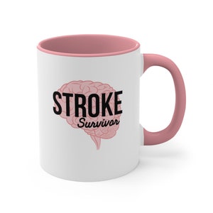 Può includere: Tazza in ceramica bianca con bordo e manico rosa. La tazza presenta una grafica di cervello rosa con il testo "STROKE Survivor" in nero.