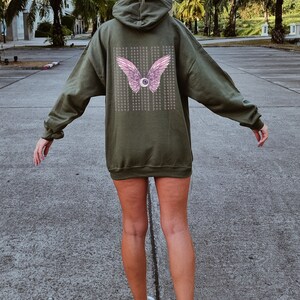 May include: A green hooded sweatshirt with a pink eye and wings graphic on the back. The graphic has the text "Funny seeing you here" repeated multiple times.