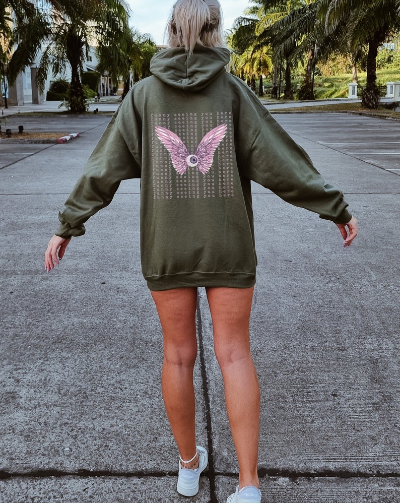 May include: A green hooded sweatshirt with a pink eye and wings graphic on the back. The graphic has the text "Funny seeing you here" repeated multiple times.