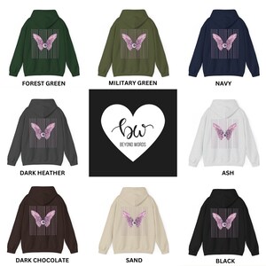 May include: A collection of hoodies in various colors, featuring a pink and white graphic of wings with an eye on the back. The hoodies are available in Forest Green, Military Green, Navy, Dark Heather, Ash, Dark Chocolate, Sand, and Black. The logo "bw BEYOND WORDS" is displayed in a white heart on a black background.