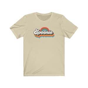 Op de afbeelding: Een beige t-shirt met een retro-regenbooggrafiek en het woord "Spoonie" in zwarte tekst.